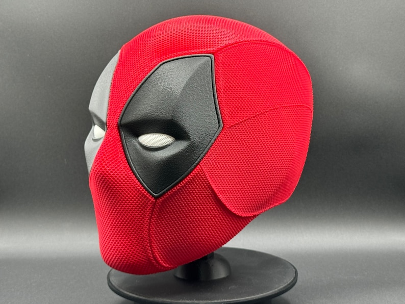 May include: A red and black Deadpool mask with a textured surface. The mask is on a black stand.