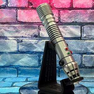 May include: A silver lightsaber prop with red and green buttons, resting on a black stand. The lightsaber has a ribbed handle and a detailed emitter. The background is a brick wall with blue, pink, and purple hues.