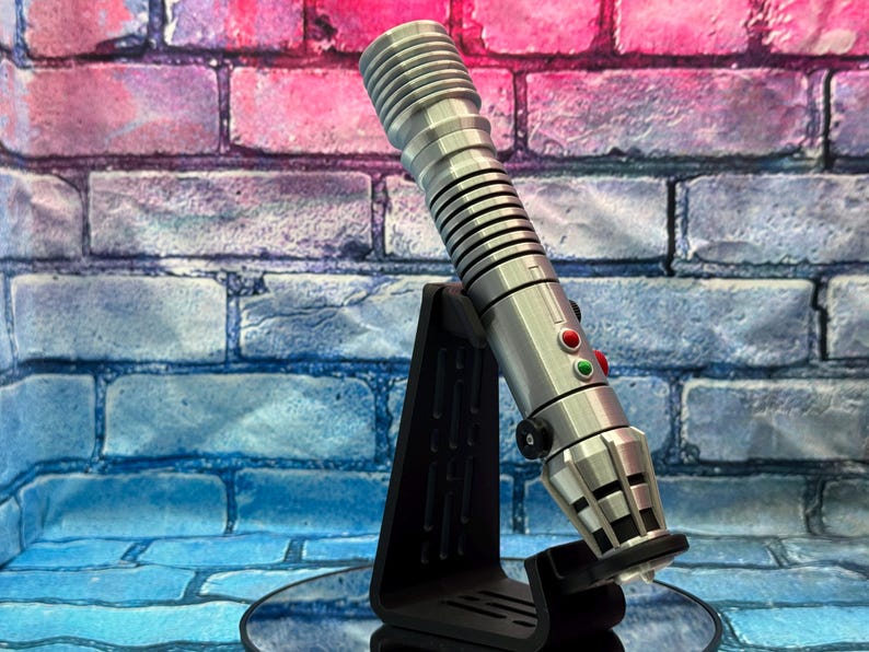 May include: A silver lightsaber prop with red and green buttons, resting on a black stand. The lightsaber has a ribbed handle and a detailed emitter. The background is a brick wall with blue, pink, and purple hues.