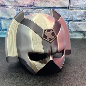 May include: A silver and black helmet with a geometric design. The helmet has a visor and ear coverings. A crest with a bird-like emblem is on the front. The helmet is likely for cosplay or costume use.