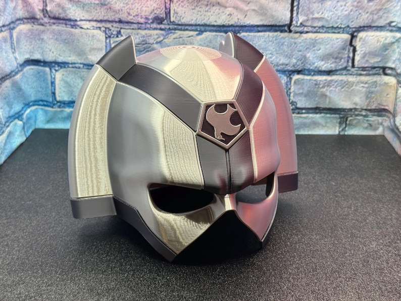 May include: A silver and black helmet with a geometric design. The helmet has a visor and ear coverings. A crest with a bird-like emblem is on the front. The helmet is likely for cosplay or costume use.