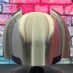 May include: A silver-colored helmet with black accents, possibly for cosplay or display. The helmet has a rounded top and angular side pieces. The background is a brick wall with pink, blue, and white paint.