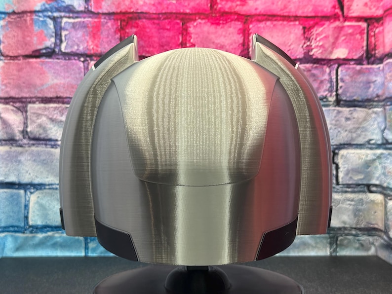 May include: A silver-colored helmet with black accents, possibly for cosplay or display. The helmet has a rounded top and angular side pieces. The background is a brick wall with pink, blue, and white paint.