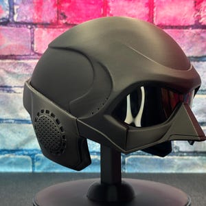 May include: A matte black helmet with a visor and integrated face guard. The helmet has a futuristic design with a circular vent on the side. It is displayed on a black stand against a brick wall background.
