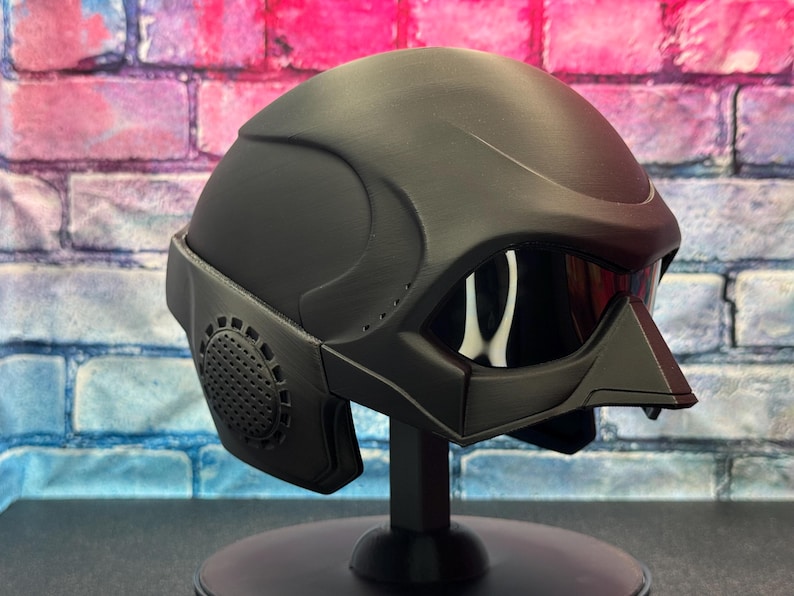 May include: A matte black helmet with a visor and integrated face guard. The helmet has a futuristic design with a circular vent on the side. It is displayed on a black stand against a brick wall background.