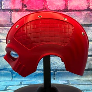 May include: A bright red, half-face mask with a textured, woven pattern on the upper portion. The mask has eye and mouth openings, and is secured with small, silver-colored rivets. The mask is displayed on a black stand against a colorful brick wall.