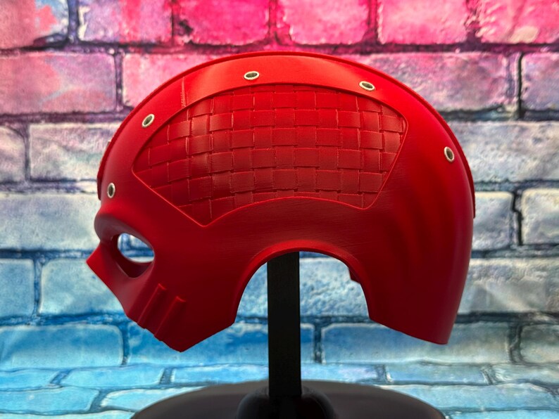 May include: A bright red, half-face mask with a textured, woven pattern on the upper portion. The mask has eye and mouth openings, and is secured with small, silver-colored rivets. The mask is displayed on a black stand against a colorful brick wall.