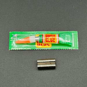 Puede incluir: Un paquete verde y blanco de superpegamento con un tubo de pegamento naranja y blanco en su interior. El paquete dice "Super Glue" y "World Class Quality First Choice". Tambi&eacute;n hay una pila de peque&ntilde;os imanes redondos plateados.