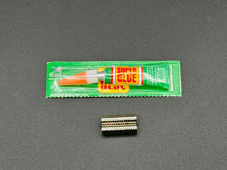 Puede incluir: Un paquete verde y blanco de superpegamento con un tubo de pegamento naranja y blanco en su interior. El paquete dice "Super Glue" y "World Class Quality First Choice". Tambi&eacute;n hay una pila de peque&ntilde;os imanes redondos plateados.