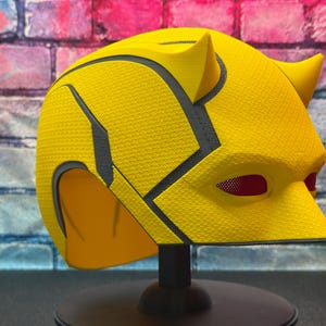 Puede incluir: Un casco de superh&eacute;roe amarillo con una superficie texturizada y detalles en negro. El casco presenta dos peque&ntilde;os cuernos y cubiertas oculares de malla roja. Se exhibe sobre un soporte negro contra una pared de ladrillos.
