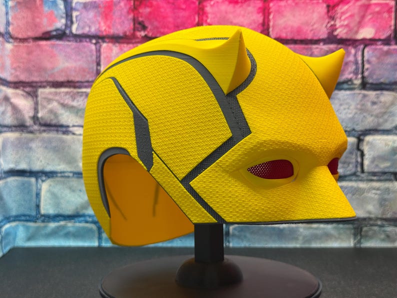 Puede incluir: Un casco de superh&eacute;roe amarillo con una superficie texturizada y detalles en negro. El casco presenta dos peque&ntilde;os cuernos y cubiertas oculares de malla roja. Se exhibe sobre un soporte negro contra una pared de ladrillos.