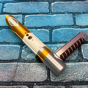 May include: A gold, white, and silver lightsaber hilt with a second, brown and silver hilt. The lightsaber has a sleek design with a metallic finish. The hilts are set against a blue brick background.