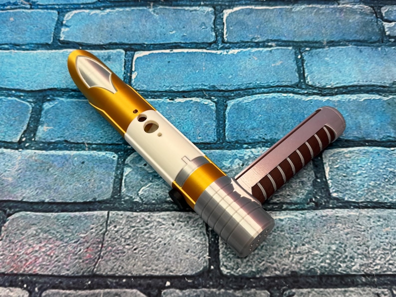 May include: A gold, white, and silver lightsaber hilt with a second, brown and silver hilt. The lightsaber has a sleek design with a metallic finish. The hilts are set against a blue brick background.