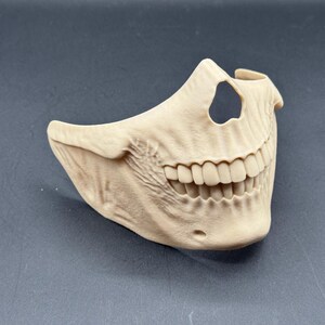 Puede incluir: Una m&aacute;scara de calavera beige, que cubre la mitad inferior de la cara, con dientes y mand&iacute;bula detallados. La m&aacute;scara tiene un recorte encima de los dientes. Est&aacute; hecha de un material s&oacute;lido, probablemente para disfraz o decoraci&oacute;n.