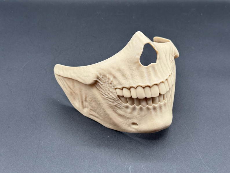 Puede incluir: Una m&aacute;scara de calavera beige, que cubre la mitad inferior de la cara, con dientes y mand&iacute;bula detallados. La m&aacute;scara tiene un recorte encima de los dientes. Est&aacute; hecha de un material s&oacute;lido, probablemente para disfraz o decoraci&oacute;n.