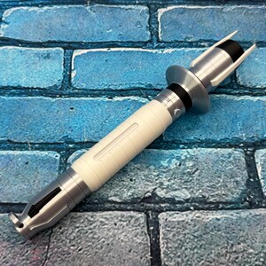 May include: A silver and white lightsaber prop with a black accent. The hilt is white with silver detailing and a black section near the top. The lightsaber is set against a blue brick background.