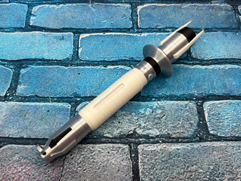 May include: A silver and white lightsaber prop with a black accent. The hilt is white with silver detailing and a black section near the top. The lightsaber is set against a blue brick background.
