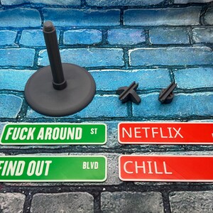 May include: A collection of novelty street signs with phrases like "FUCK AROUND ST", "FIND OUT BLVD", "NETFLIX PL", and "CHILL ST". Also included are a gray stand and two small, dark gray pieces. The signs are green and red.