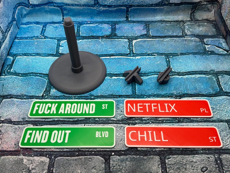 May include: A collection of novelty street signs with phrases like "FUCK AROUND ST", "FIND OUT BLVD", "NETFLIX PL", and "CHILL ST". Also included are a gray stand and two small, dark gray pieces. The signs are green and red.