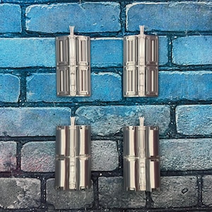 以下が含まれることがあります： 詳細なデザインの銀色の長方形のオブジェクトが4つ、グリッド状に配置されています。各オブジェクトの上部には小さな垂直のハンドルが付いています。オブジェクトは、青とグレーのレンガの壁を背景にしています。