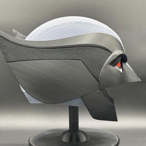 May include: A black and gray 3D printed helmet with red eyes. The helmet is on a black stand.