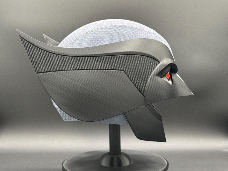 May include: A black and gray 3D printed helmet with red eyes. The helmet is on a black stand.
