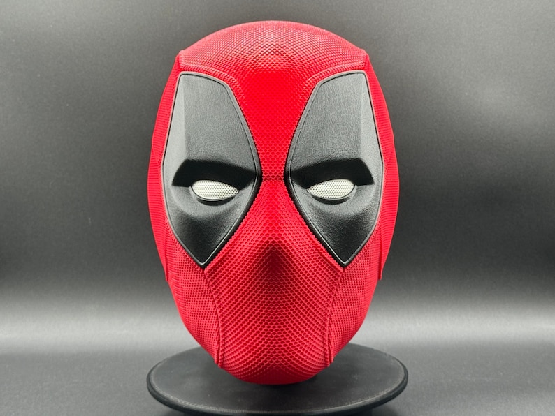 May include: A red and black Deadpool mask with a textured surface. The mask has a white eye mesh and a black eye outline.