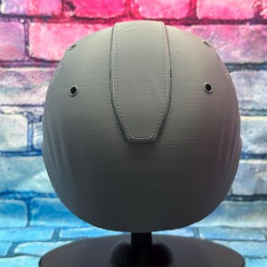 May include: A gray, spherical object with a textured surface and a stitched design on top. The object has several small, evenly spaced holes and is mounted on a black stand. The background features a brick wall with blue, pink, and purple hues.
