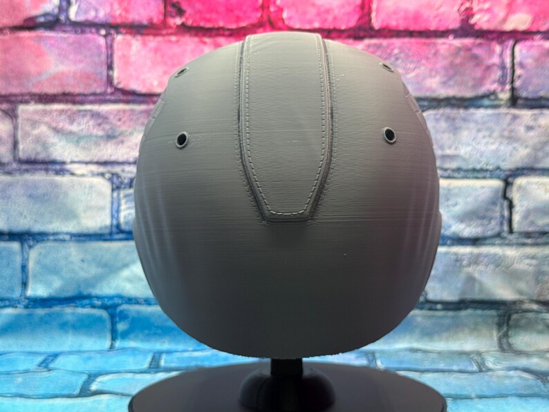 May include: A gray, spherical object with a textured surface and a stitched design on top. The object has several small, evenly spaced holes and is mounted on a black stand. The background features a brick wall with blue, pink, and purple hues.