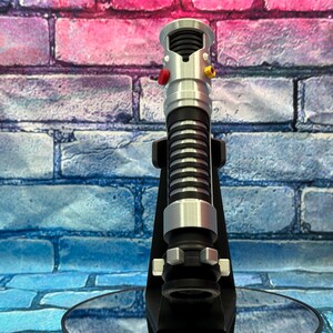 May include: A silver and black lightsaber prop with red and yellow buttons, resting on a black stand. The lightsaber has a ribbed black grip and silver accents. The background is a brick wall with blue, pink, and purple hues.