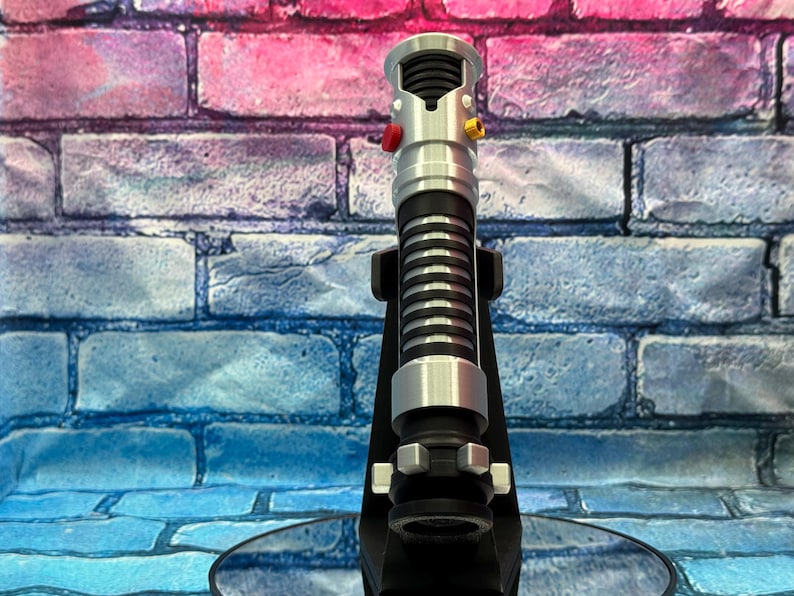May include: A silver and black lightsaber prop with red and yellow buttons, resting on a black stand. The lightsaber has a ribbed black grip and silver accents. The background is a brick wall with blue, pink, and purple hues.