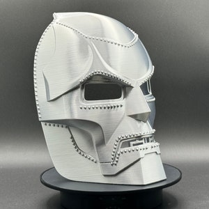 May include: A silver-colored 3D printed mask with a detailed design, resembling a character from a comic book or science fiction movie. The mask has a textured surface and a raised design around the eyes and mouth.