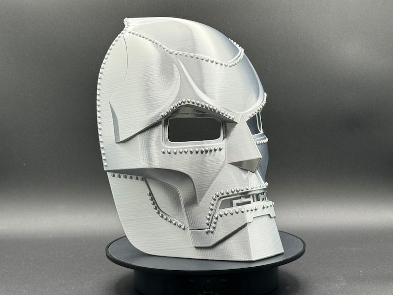 May include: A silver-colored 3D printed mask with a detailed design, resembling a character from a comic book or science fiction movie. The mask has a textured surface and a raised design around the eyes and mouth.