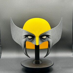 Puede incluir: Una m&aacute;scara de Wolverine, un personaje de superh&eacute;roe popular, impresa en 3D en amarillo y negro, sobre un soporte negro.
