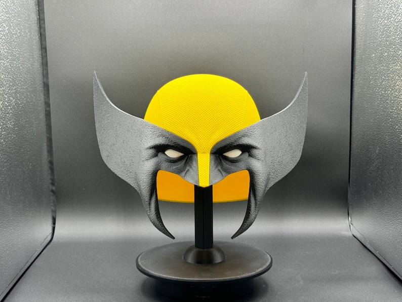 Puede incluir: Una m&aacute;scara de Wolverine, un personaje de superh&eacute;roe popular, impresa en 3D en amarillo y negro, sobre un soporte negro.