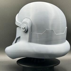 May include: A white 3D printed helmet with a textured surface. The helmet has a rounded top and a curved visor. The helmet is sitting on a black base.