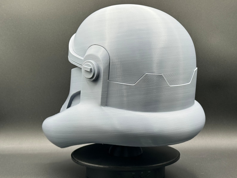 May include: A white 3D printed helmet with a textured surface. The helmet has a rounded top and a curved visor. The helmet is sitting on a black base.