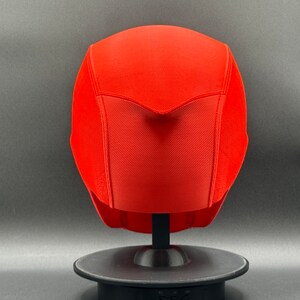 K&ouml;nnte beinhalten: Ein leuchtend roter Helm mit einer strukturierten Oberfl&auml;che und einer spitzen Oberseite. Der Helm wird auf einem schwarzen St&auml;nder pr&auml;sentiert.