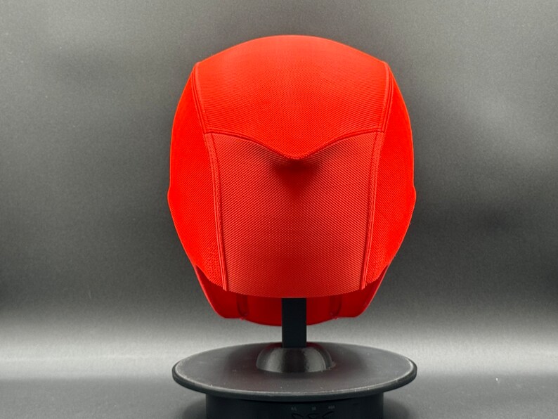 K&ouml;nnte beinhalten: Ein leuchtend roter Helm mit einer strukturierten Oberfl&auml;che und einer spitzen Oberseite. Der Helm wird auf einem schwarzen St&auml;nder pr&auml;sentiert.