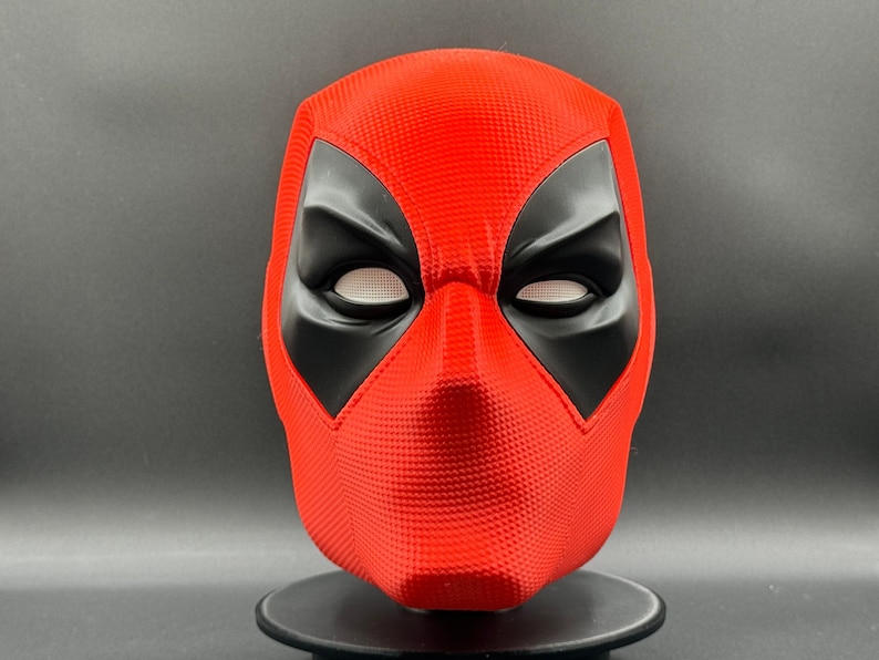 Puede incluir: Una m&aacute;scara de Deadpool roja y negra con una superficie texturizada. La m&aacute;scara tiene ojos blancos y una boca negra.