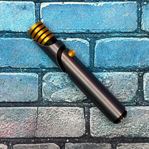 May include: A metallic gray lightsaber with gold accents and a gold button. The hilt has a black grip and a gold-colored emitter. The lightsaber is set against a blue brick wall.