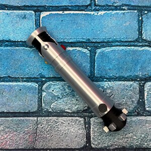 May include: A silver and black lightsaber prop with a red button and yellow button. The prop is lying diagonally on a blue brick wall background. The lightsaber has a cylindrical body and a detailed hilt.