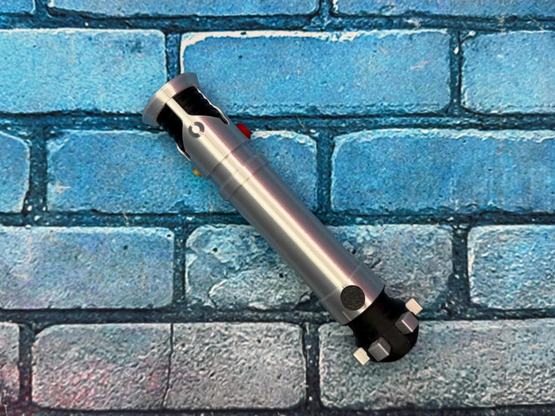 May include: A silver and black lightsaber prop with a red button and yellow button. The prop is lying diagonally on a blue brick wall background. The lightsaber has a cylindrical body and a detailed hilt.