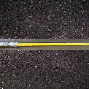 May include: A yellow lightsaber with a silver handle and red and green buttons. The lightsaber is set against a starry background, evoking a space theme. The blade is long and straight, and the handle has intricate details.