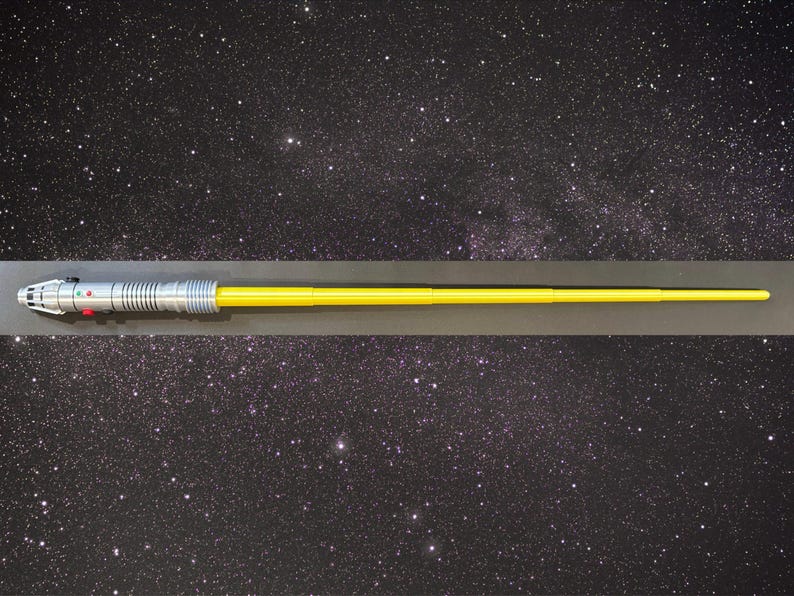 May include: A yellow lightsaber with a silver handle and red and green buttons. The lightsaber is set against a starry background, evoking a space theme. The blade is long and straight, and the handle has intricate details.