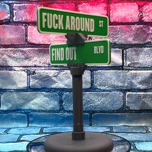 May include: A novelty street sign with green and white lettering. The top sign reads "FUCK AROUND ST" and the bottom sign reads "FIND OUT BLVD". The sign is mounted on a gray post with a round base, set against a brick wall.