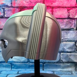 May include: A silver-colored helmet with a light green accent on top. The helmet has a detailed design with vertical lines on the side and a face opening. It is displayed on a black stand against a brick wall.
