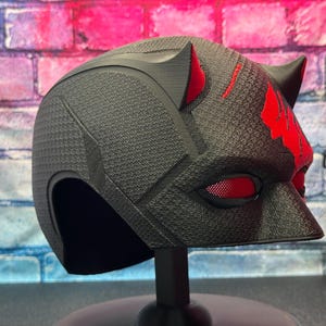 May include: A black helmet with red accents, featuring a textured surface and cat-like ears. The eye openings have a red mesh. The helmet is displayed on a black stand, against a brick wall background.
