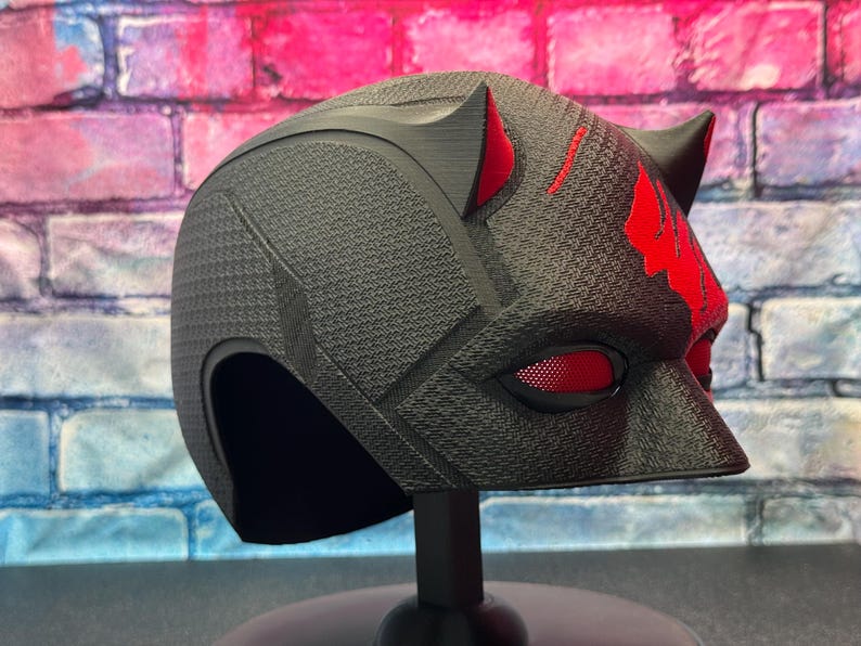 May include: A black helmet with red accents, featuring a textured surface and cat-like ears. The eye openings have a red mesh. The helmet is displayed on a black stand, against a brick wall background.