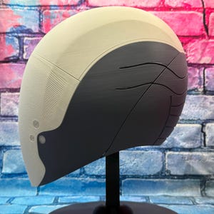 May include: A futuristic-looking helmet with a two-tone design. The top portion is off-white, while the lower section is dark gray with curved etched lines. The helmet is mounted on a black stand, set against a brick wall.